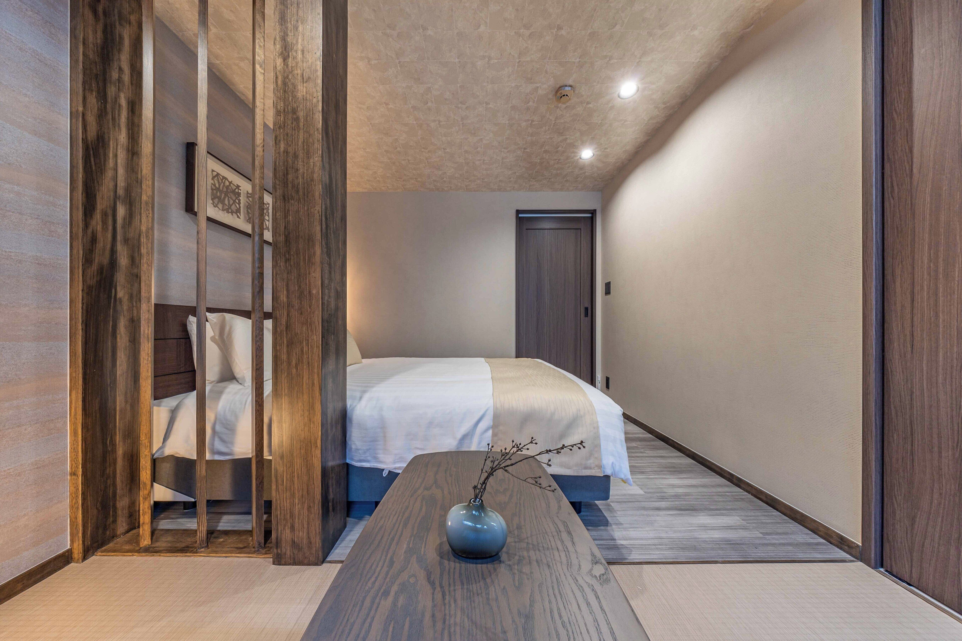 A hotel room with a bed, a wooden partition, a vase with branches, and a wooden floor.