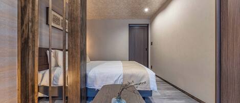A hotel room with a bed, a wooden partition, a vase with branches, and a wooden floor.