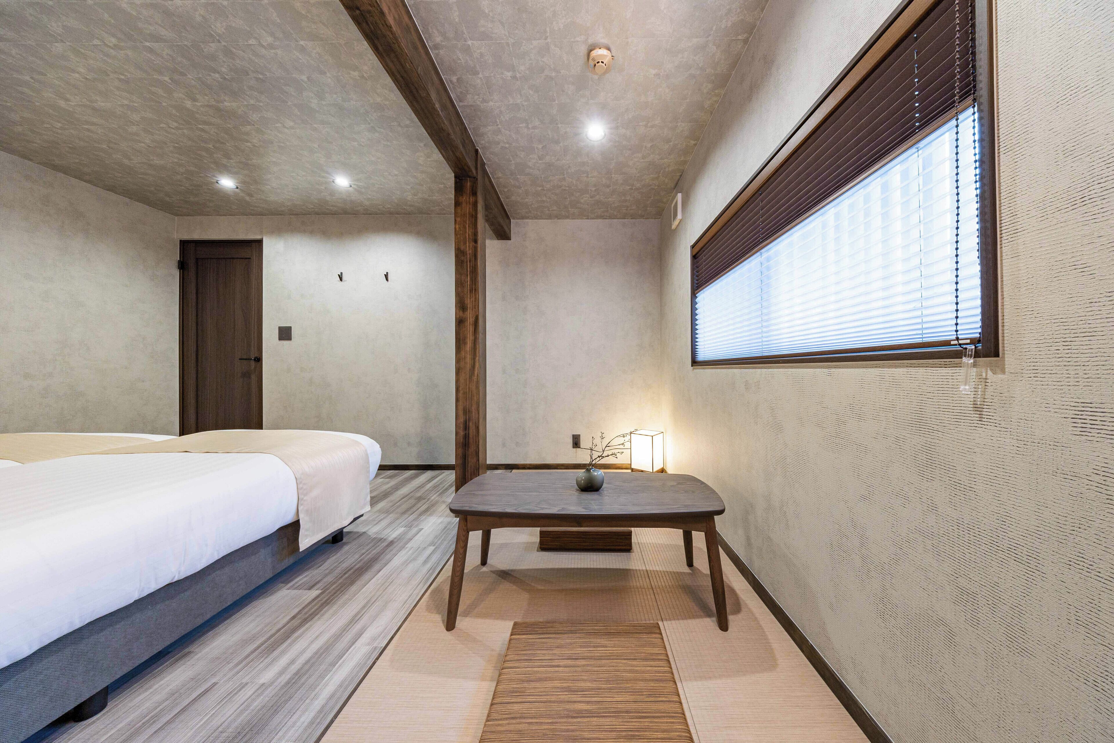 A hotel room with a bed, a small table with a vase, and a window with blinds.