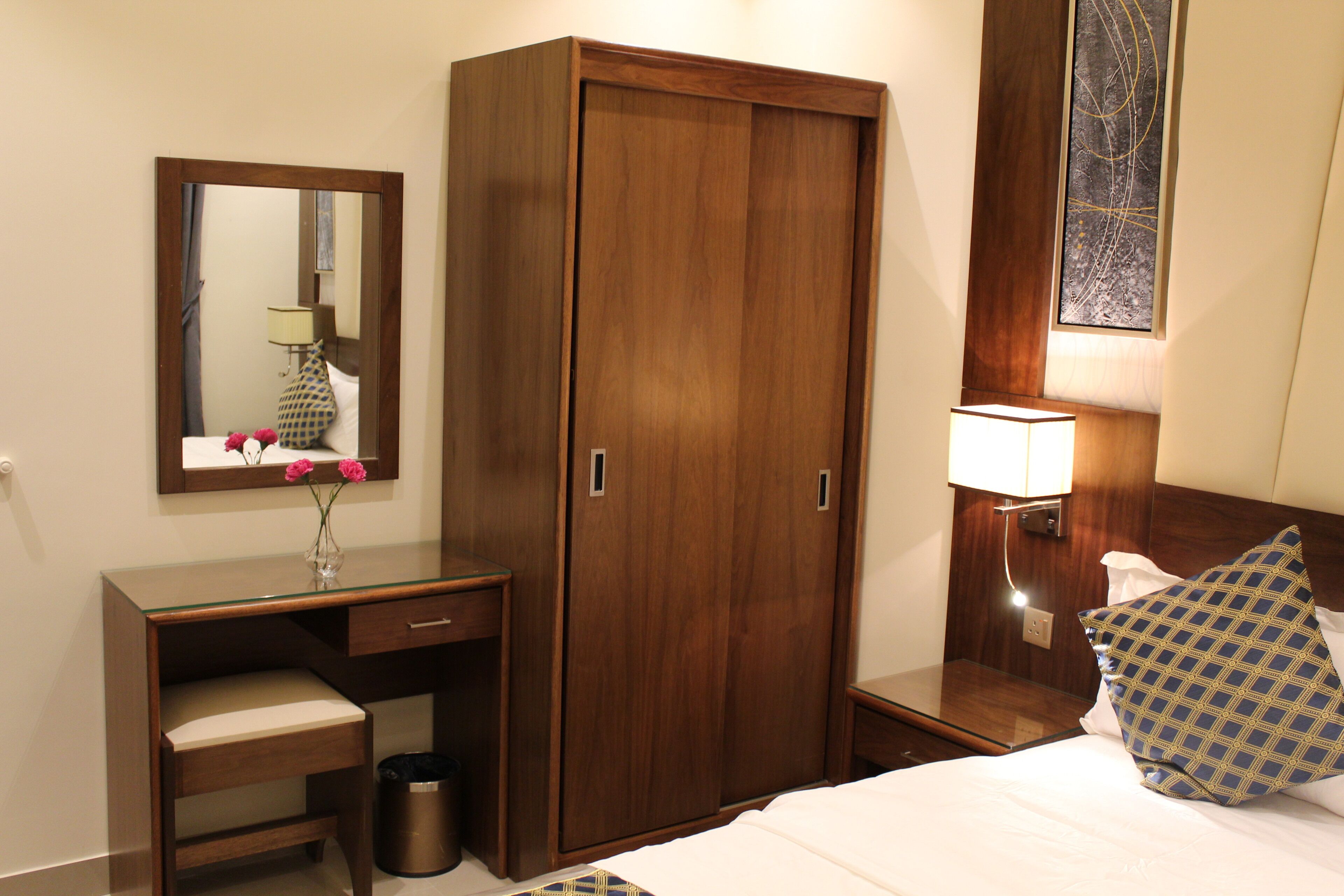 A hotel room with a wooden wardrobe, a desk with a mirror, a bedside table with a lamp, and a bed with patterned pillows.