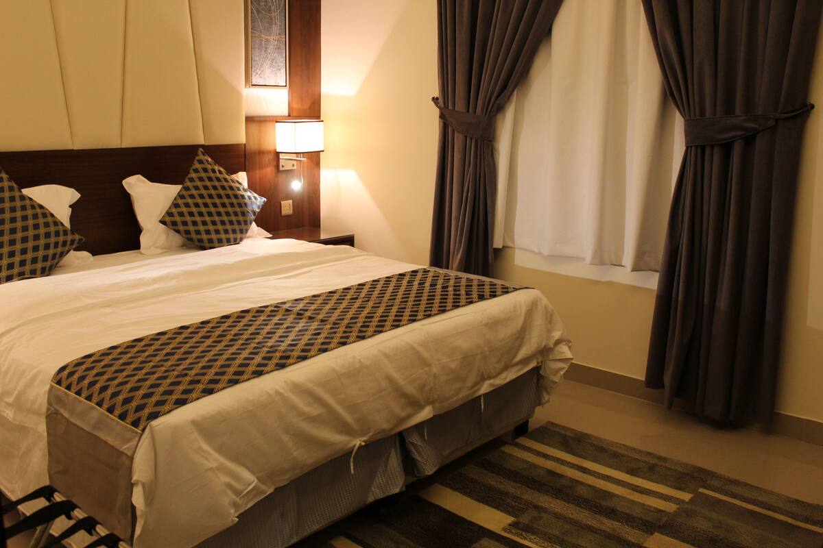 Habitación de hotel con una cama grande, cabecero, mesita de noche con lámpara y cortinas.