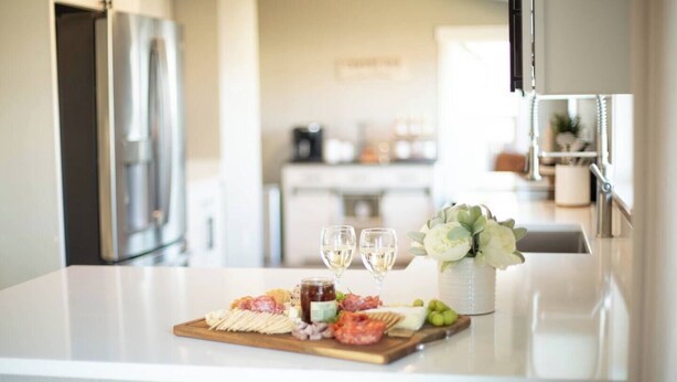 A kitchen counter with a charcuterie board, two glasses of white wine, and a vase with flowers.