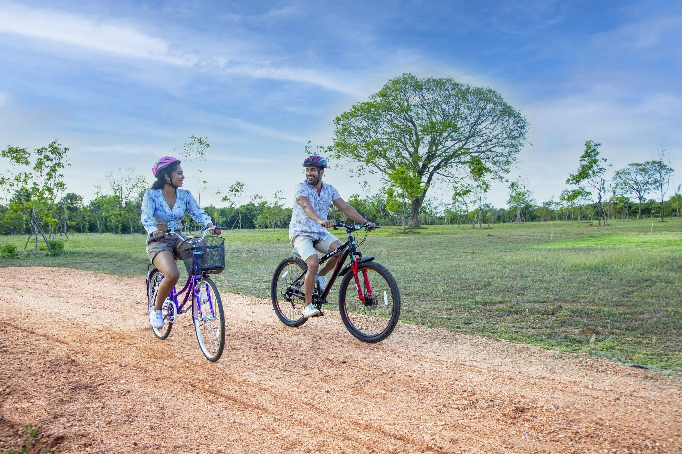 Dos personas montando en bicicleta por un sendero de tierra, con árboles y césped al fondo.