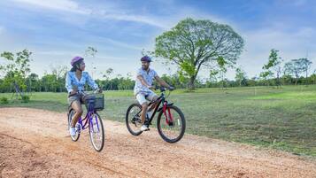 Two people riding bicycles on a dirt path with trees and grass in the background.