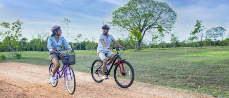 Two people riding bicycles on a dirt path with trees and grass in the background.