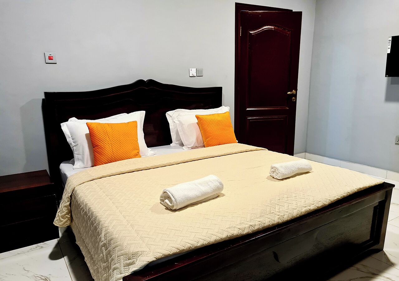 A neatly made bed with a beige quilt, two white towels, and two orange pillows.