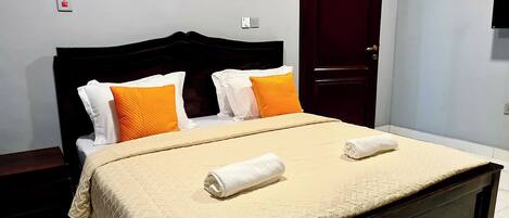 A neatly made bed with a beige quilt, two white towels, and two orange pillows.