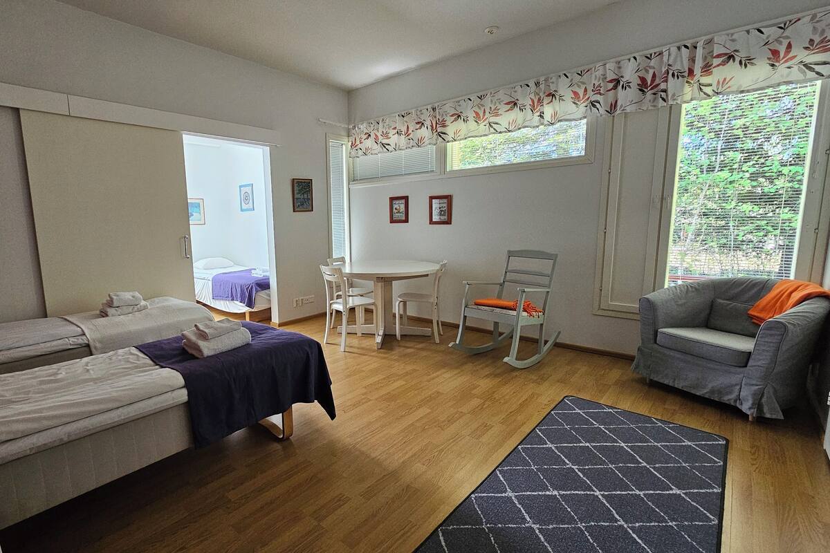 Una habitación con dos camas, una mesa de comedor con sillas, un sillón mecedor, un sofá y una ventana con persianas.