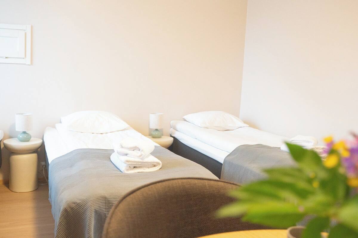 Habitación de hotel con dos camas individuales, cada una con una almohada y una toalla enrollada. Mesitas de noche con lámparas y un pequeño florero sobre una de ellas.