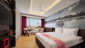Quarto de hotel moderno com uma cama grande, televisão de tela plana, área de refeições com mesa e cadeiras, e um mural com vista para um rio e montanhas.