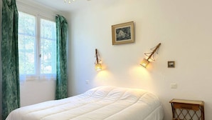 Ein Schlafzimmer mit Bett, Fenster mit Vorhängen, einer Wandlampe und einem kleinen Tisch.