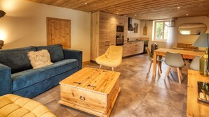 A modern interior with a wooden ceiling, a blue sofa, a yellow armchair, a wooden chest, and a dining area with chairs and a table. - Renovated apartment in central Morillon for 6 ppl. (Morillon Village)