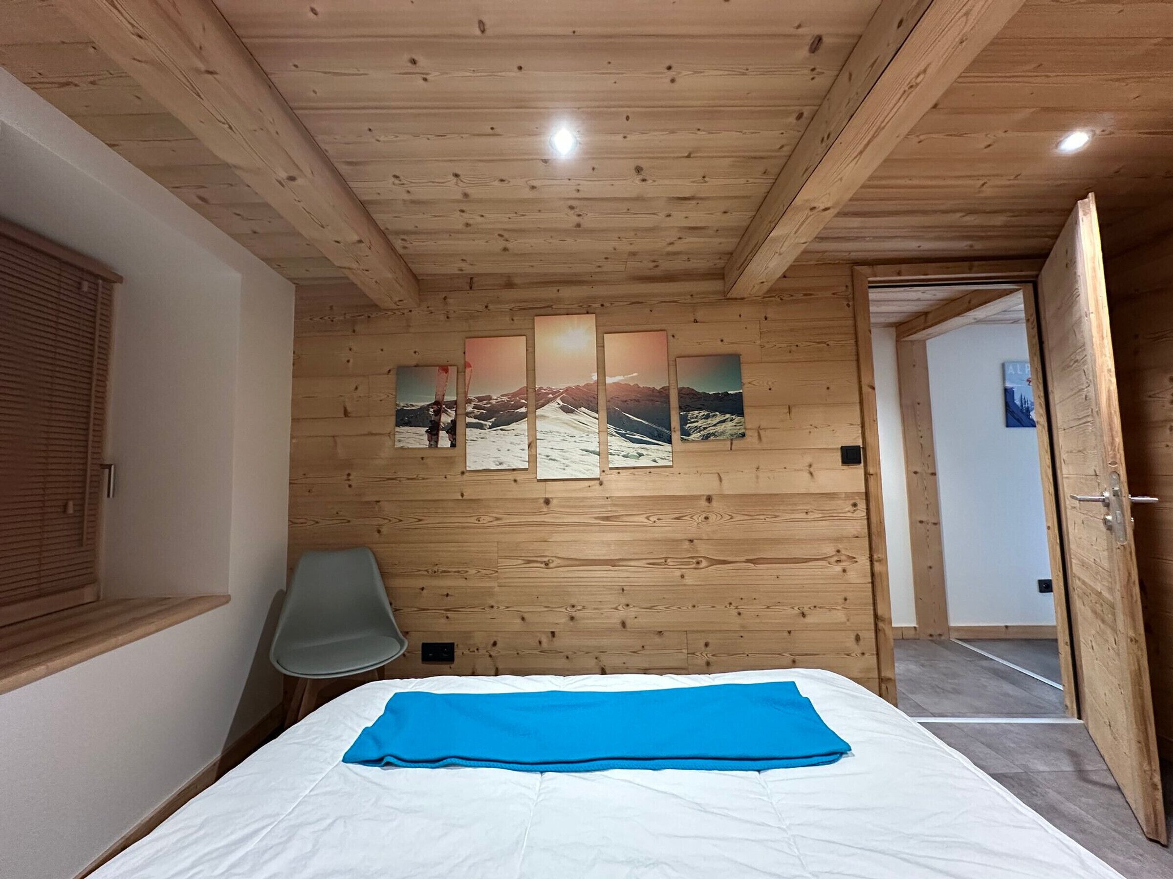 Una habitación con paneles de madera, una cama con manta azul, una silla y una puerta que da a otra habitación.