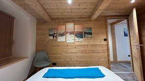 A room with wooden panel walls, a bed with a blue blanket, a chair, and a door leading to another room. - Renovated apartment in central Morillon for 6 ppl. (Morillon Village)