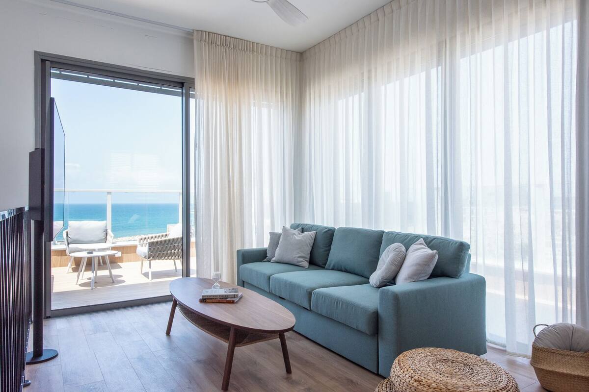 A modern living room with a teal sofa, a wooden coffee table, and a view of the ocean through large windows.