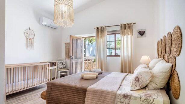 A bedroom with a large bed, a crib, a hanging light fixture, and a view of the outdoors. - Traditional Algarve home with private pool, air con & solar, near Praia Marinha (Lagoa)