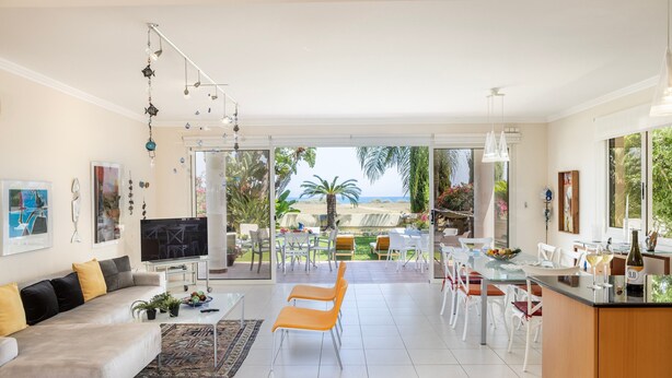 A modern living room with a sofa, TV, and dining area with chairs and a table. - Larnaca Chelidonia Seafront Villa (Pervolia)