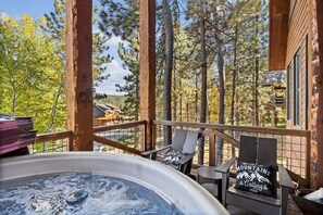 A hot tub on a wooden deck with chairs, surrounded by trees and a mountain view.