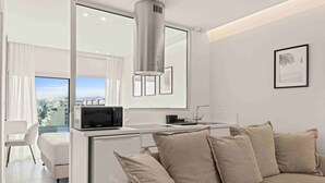 Un espacio habitable moderno y compacto con kitchenette, un sofá con cojines, una zona de comedor y un amplio ventanal con vistas a la ciudad.