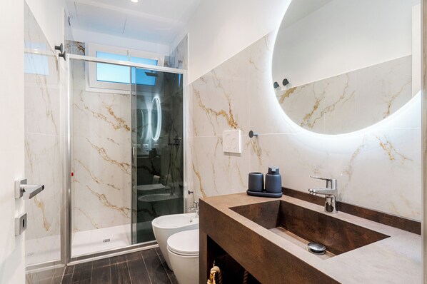 A modern bathroom with a glass shower enclosure, a marble-tiled wall, a round mirror, and a dark brown sink.