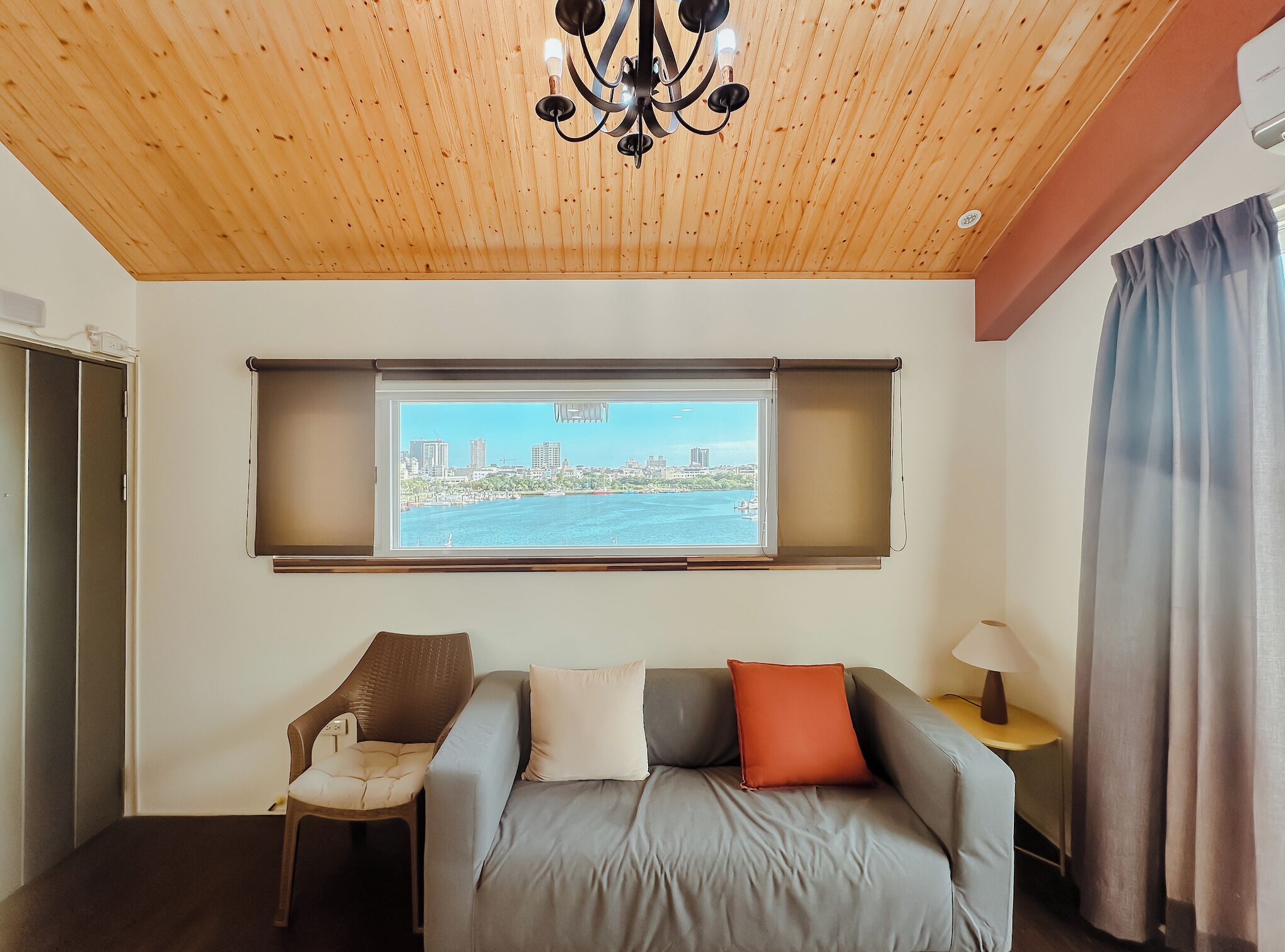 A room with a wooden ceiling, a sofa with cushions, a chair, a window with a view of a cityscape, and a lamp on a side table.