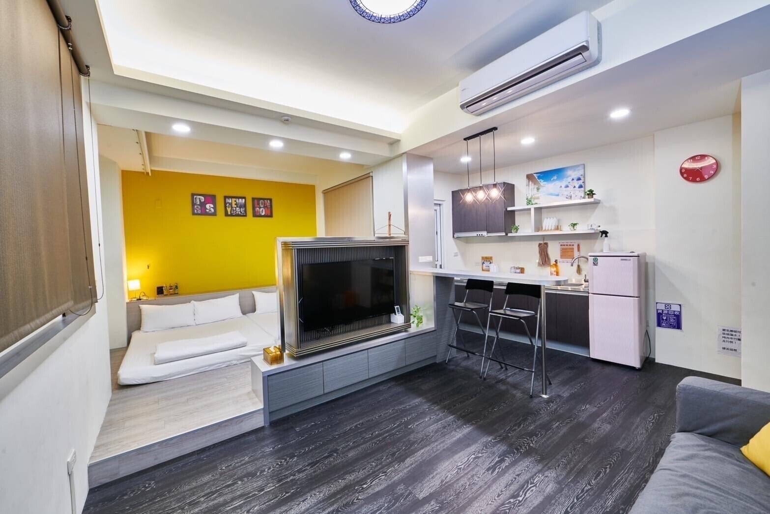 A compact hotel room with a sofa bed, a TV, a kitchenette, and a small seating area with a yellow wall.