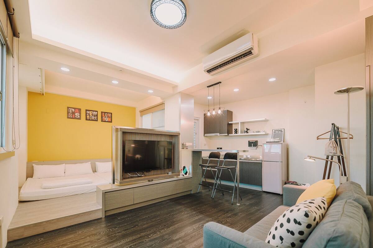 A modern living room with a flat-screen TV, a sofa, and a dining area with a kitchen in the background.