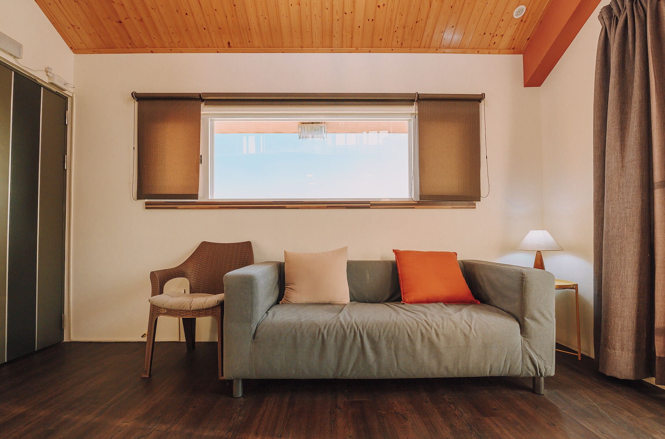 A room with a wooden ceiling, a grey sofa with cushions, a wicker chair, a window with blinds, a lamp on a side table, and a door.