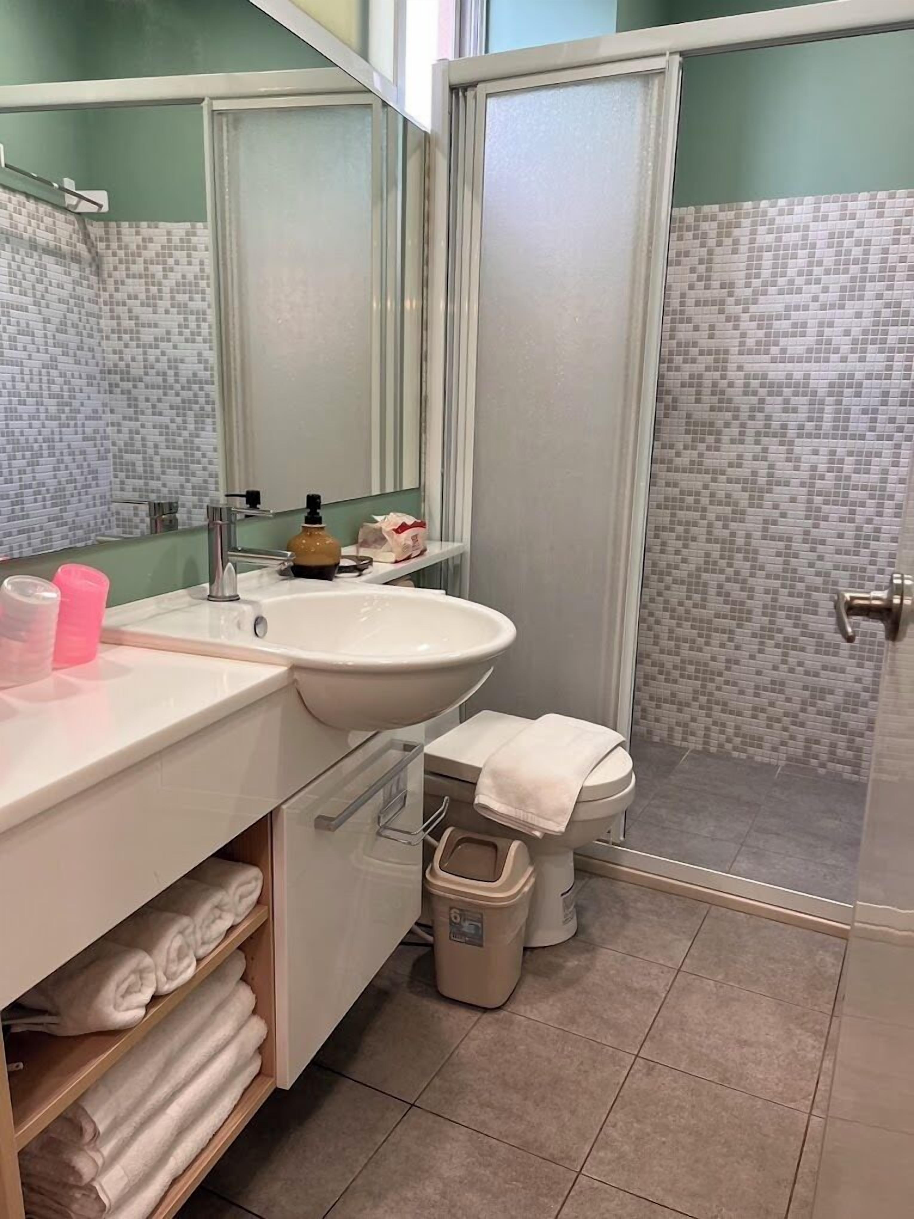 Une salle de bain avec un lavabo blanc, un miroir et des toilettes. On y trouve un porte-serviettes avec des serviettes pliées et une poubelle.