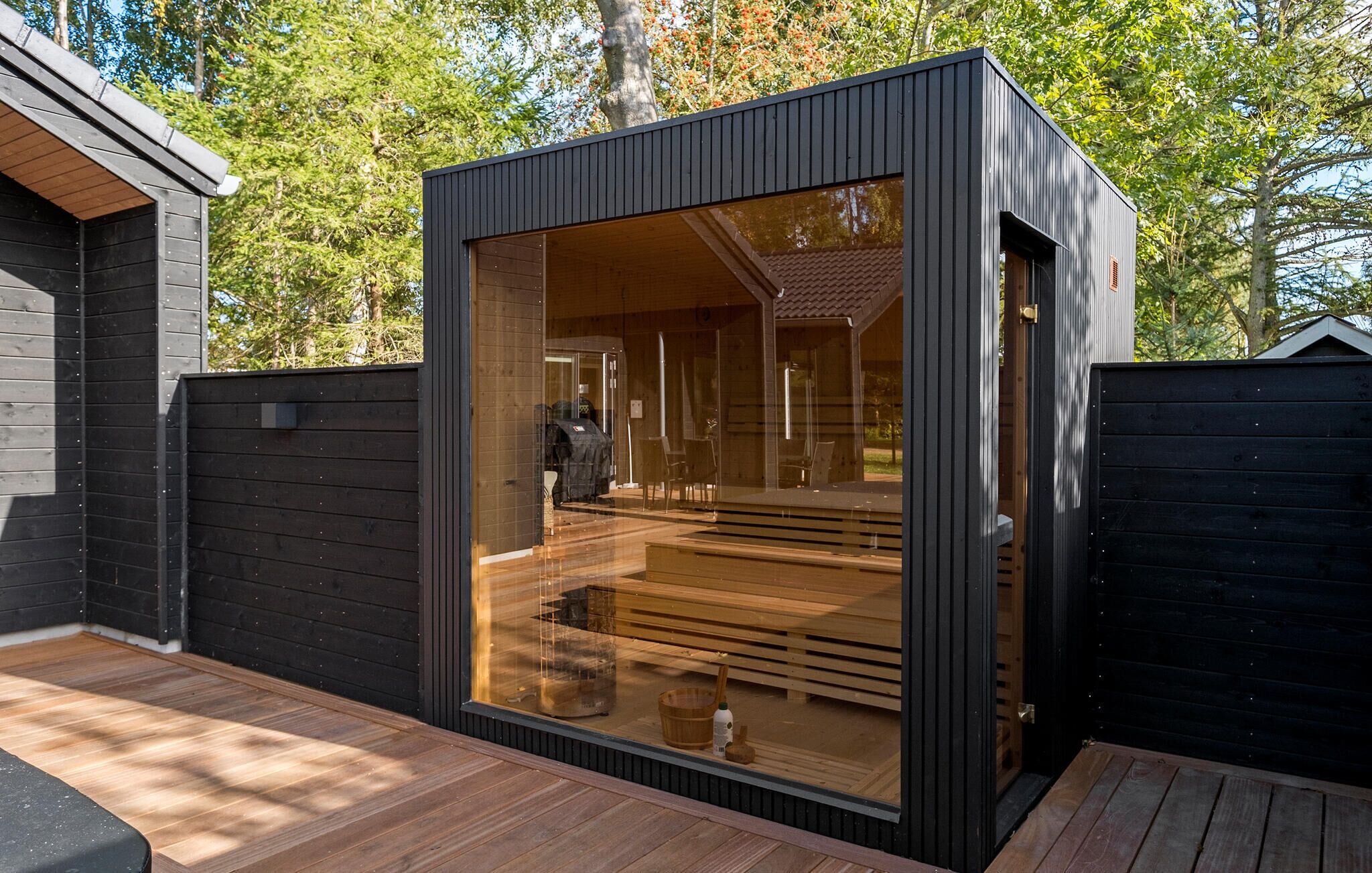 A modern, compact wooden cabin with a large glass window, wooden deck, and a view of the surrounding trees.
