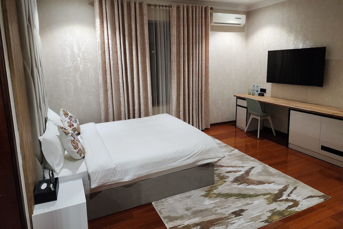 A hotel room with a bed, a desk, a chair, a TV, and an air conditioning unit.