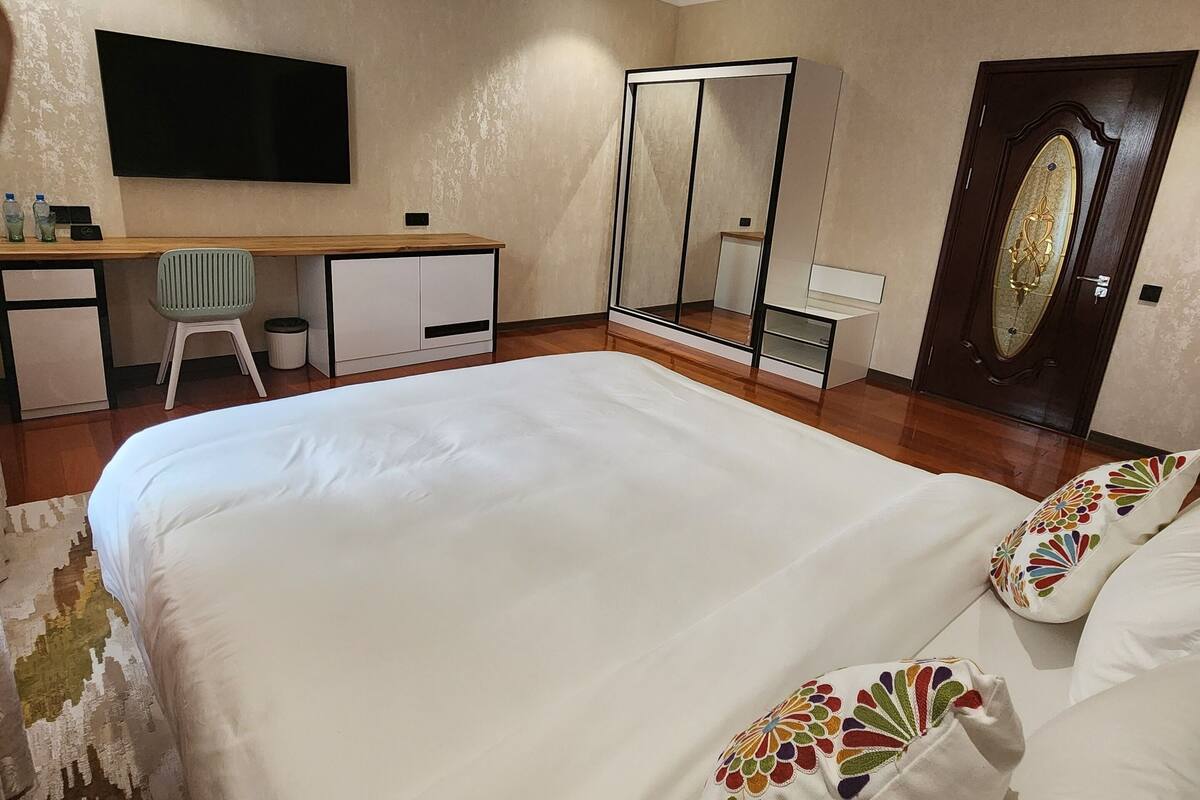 A bedroom with a large bed, a desk, a chair, a TV, and a wardrobe.
