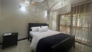 A bedroom with a bed, a nightstand, a window with curtains, and a ceiling light fixture. - Montane Safaris Hotel (Fort Portal)