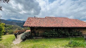 Edificio de piedra con techo de tejas, rodeado de vegetación y colinas.