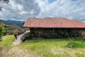 Edificio de piedra con techo de tejas, rodeado de vegetación y colinas.