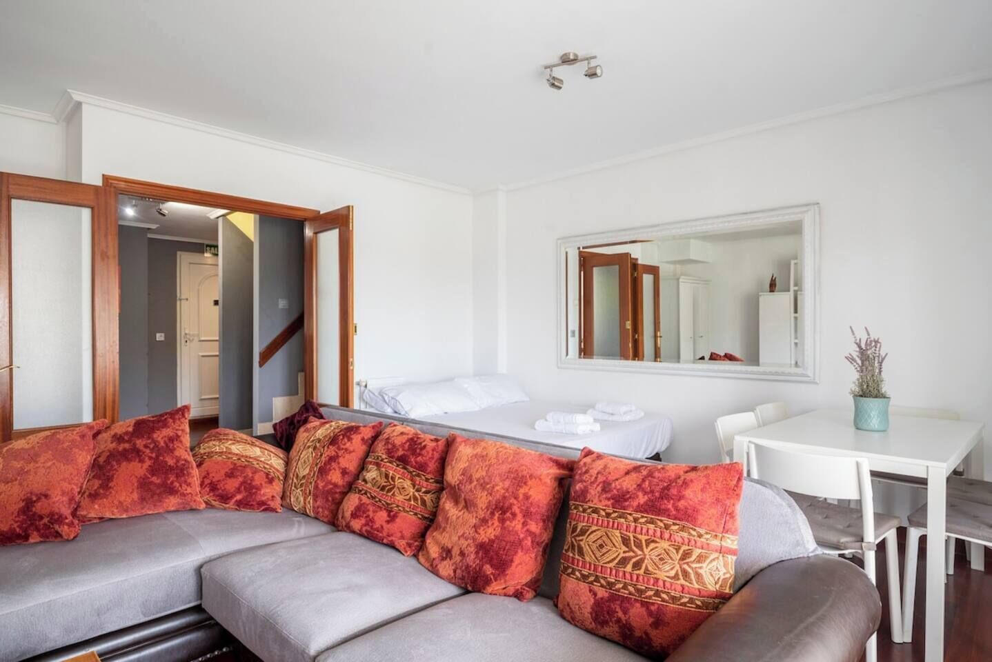 Una habitación moderna con un sofá gris, una cama blanca, una mesa de comedor y un espejo grande en la pared.