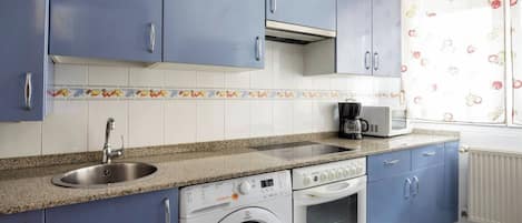 Een keuken met blauwe kastjes, een roestvrijstalen spoelbak, een koffiezetapparaat, een wasmachine en een magnetron.