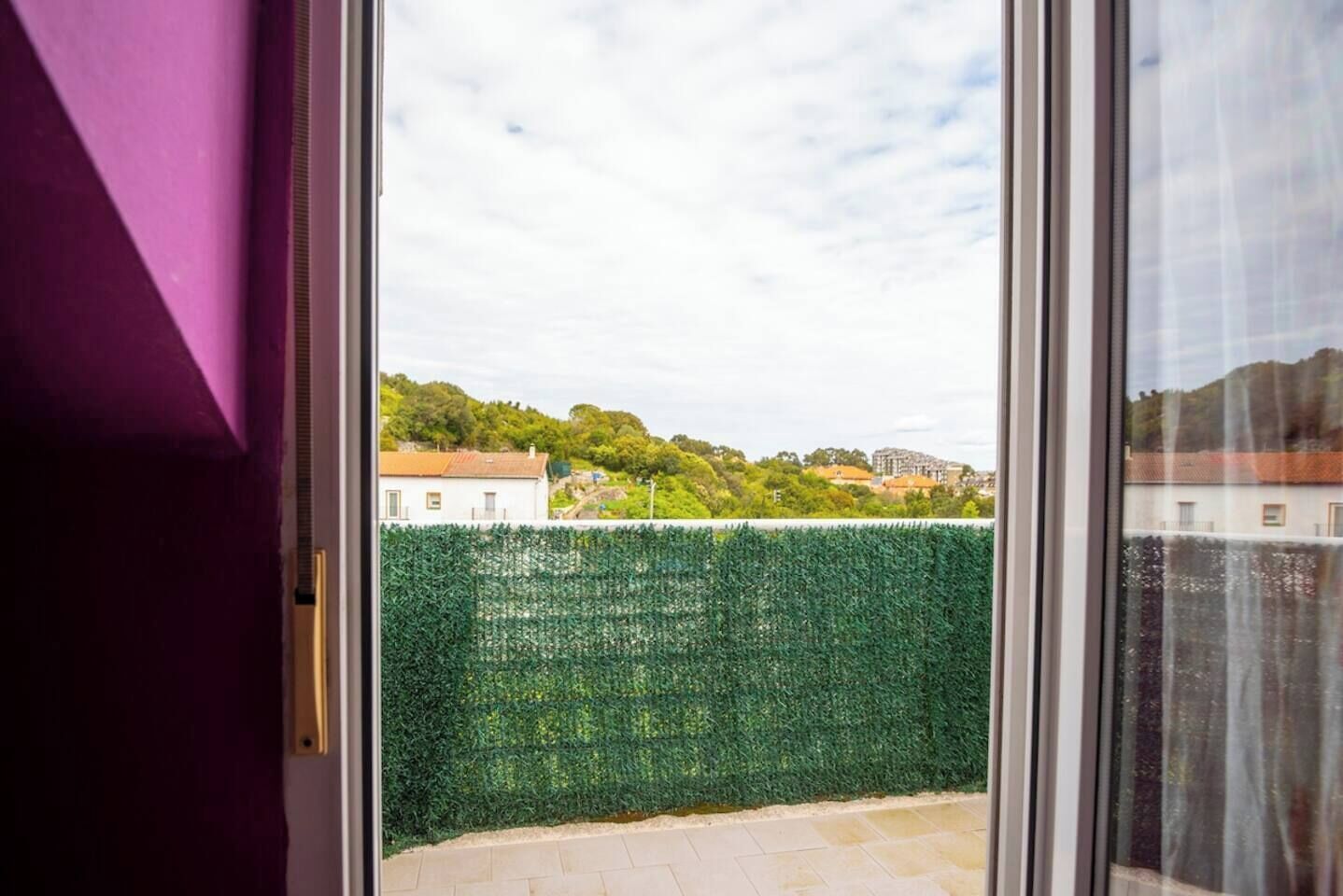 Vista a través de una ventana que muestra un seto verde, un edificio blanco y un cielo despejado.
