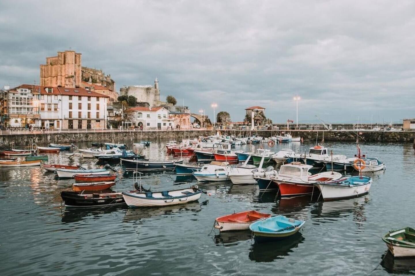 A harbor with numerous boats docked, a historic building in the background, and a calm water surface.