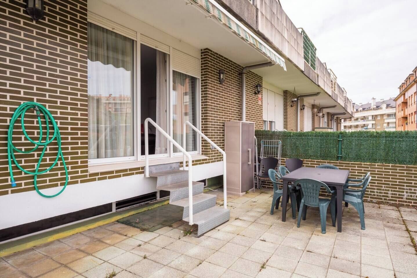 En boligbygning med en patio, hvor der er et bord, stole, en haveslange og en trappe til en øvre etage.