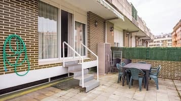En boligbygning med en patio, hvor der er et bord, stole, en haveslange og en trappe til en øvre etage.