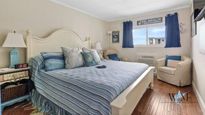 A bedroom with a bed, bedside tables, a chair, a wall clock, a sign reading "The Beach", and a view of the outside. - 1 Bedroom Ocean View Condo with Community Pool (Ocean City)