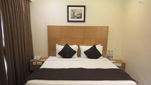 Habitación de hotel con una cama grande, dos almohadas negras, un cabecero de madera, un cuadro enmarcado en la pared, una mesita de noche con un teléfono y botellas de agua, y una cortina en la ventana.