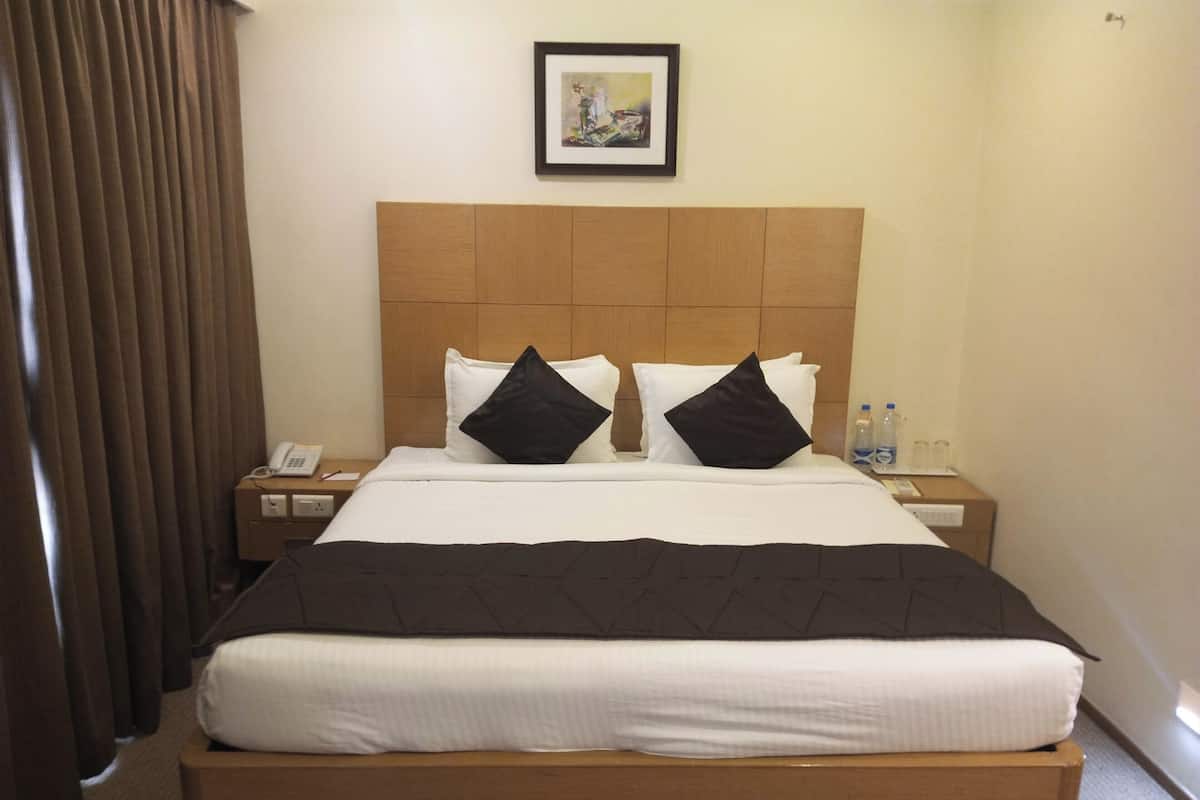 Een hotelkamer met een groot bed, twee zwarte kussens, een houten hoofdbord, een lijst met een foto aan de muur, een nachtkastje met een telefoon en waterflesjes, en een gordijn voor het raam.