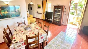 Un coin repas avec une table dressée pour quatre personnes, entourée de chaises en bois. Un téléviseur est fixé au mur, et on aperçoit un meuble contenant de la verrerie.