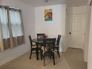 Zona de dinar amb taula rodona i quatre cadires, un quadre a la paret i una porta al fons.