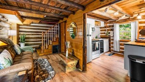 A log cabin interior with a wooden staircase, a living room with a sofa and a fur rug, a kitchen with modern appliances, and a window with curtains.