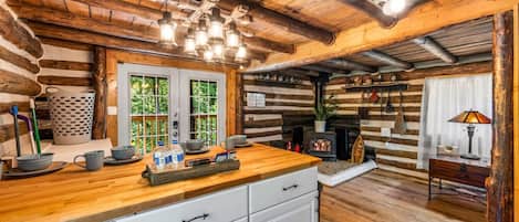 Interior rústico de una cabaña con una mesa de comedor de madera, armarios blancos, chimenea y una lámpara colgante.