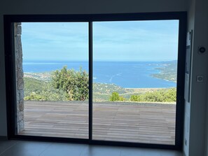A room with a large sliding glass door leading to a balcony with a view of the sea and hills.