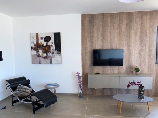 A modern living room with a black leather sofa, a round coffee table, a flat-screen TV mounted on a wooden wall, a small side table with a book, and a decorative vase with flowers.